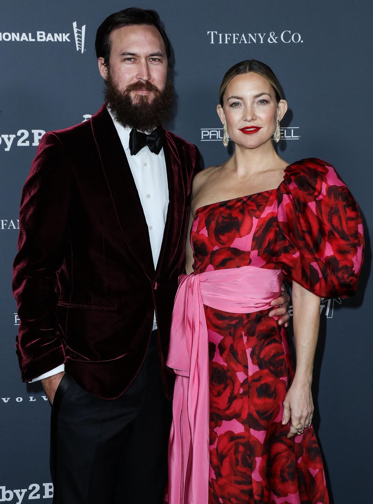 Kate Hudson stuns in a red and pink floral gown with a bold puff sleeve and waist-tied sash, standing next to fianc&eacute; Danny Fujikawa at a red carpet event