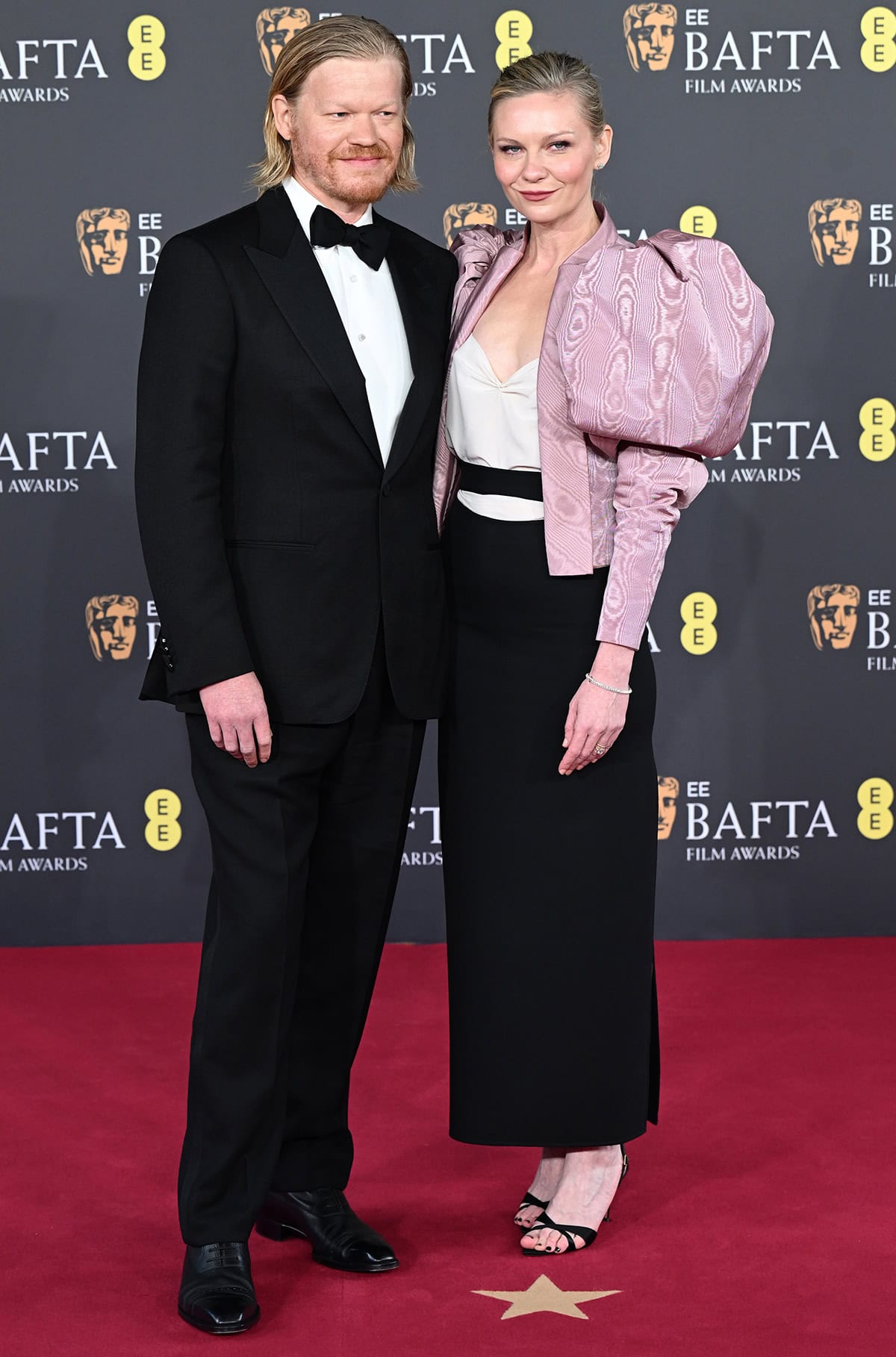 Kirsten Dunst, with her husband Jesse Plemons, power dresses in a Valentino ensemble featuring a white blouse, a black skirt, and a pink moir&eacute; jacket with oversized mutton sleeves