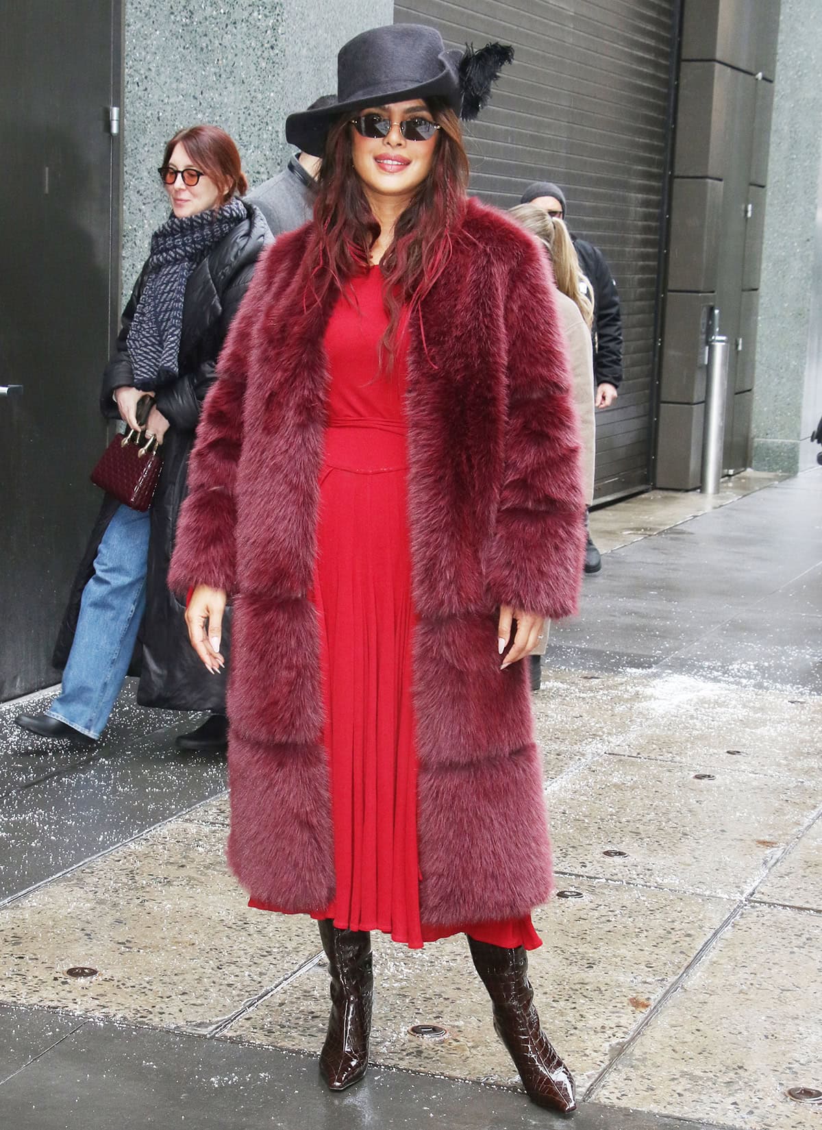 Priyanka Chopra channels piratecore energy by pairing her burgundy knee-high boots with a red jersey dress, a burgundy Apparis &ldquo;Sykle Sable&rdquo; faux-fur coat, and a black cavalier hat with a feather accent